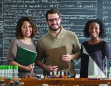 three students in front of a blackboard face a science lab
